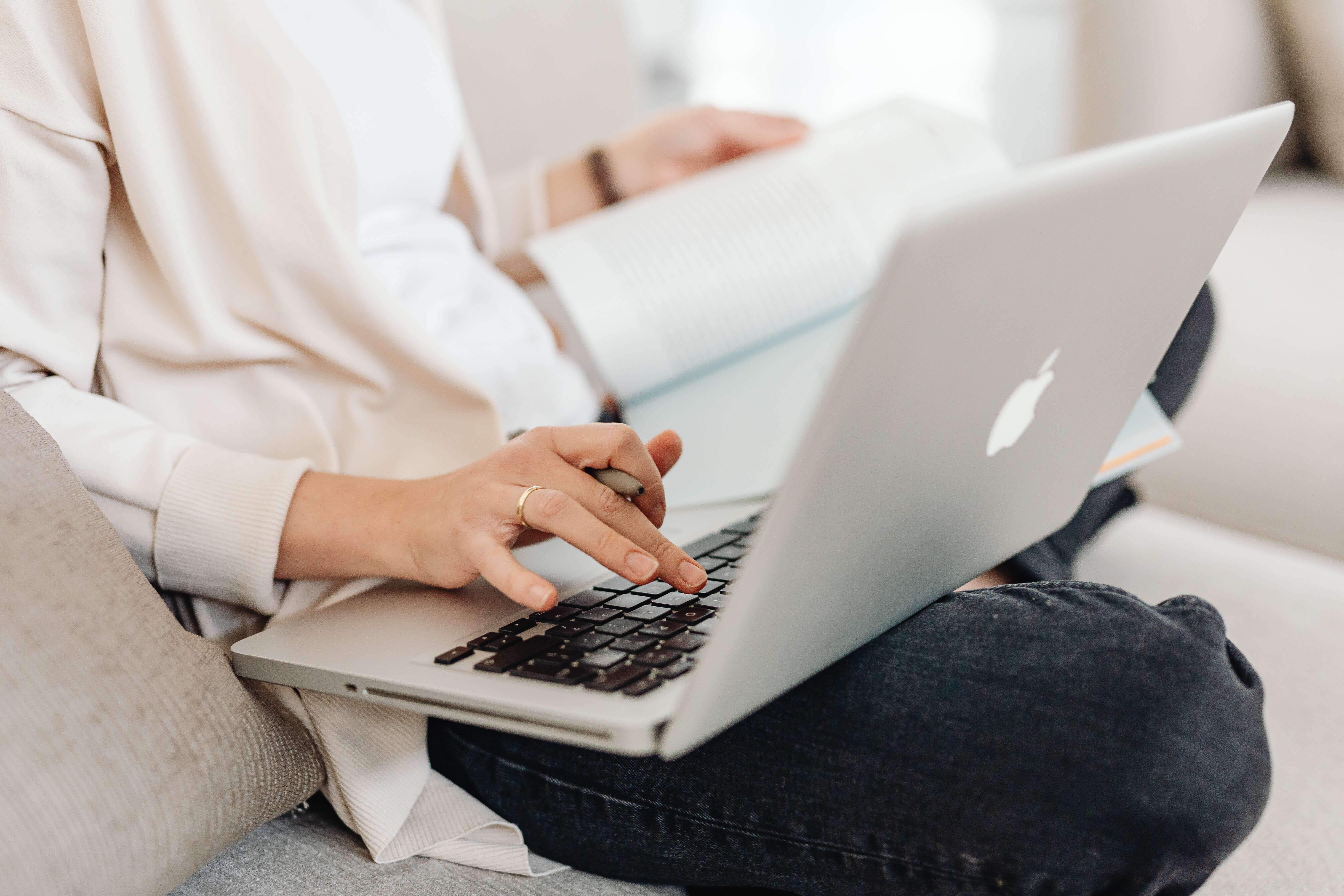 Woman's hand on a MacBook while holding a notebook in the other hand.